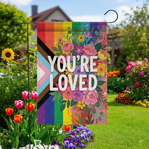 May include: A garden flag with a rainbow design and the words "YOU'RE LOVED" in white. The flag features a floral arrangement with pink, purple, and yellow flowers. A black metal garden flag holder is visible.