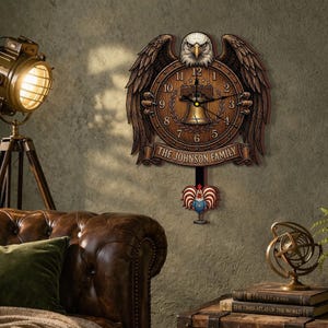 May include: A wooden wall clock featuring an eagle with outstretched wings. The clock face is brown with black numerals and hands. The text "THE JOHNSON FAMILY" is displayed below the clock face. A rooster pendulum hangs below.