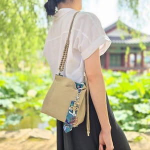 May include: A beige canvas tote bag with a long, knotted strap. The bag has a floral patterned fabric lining and a small, decorative charm hanging from the zipper pull.