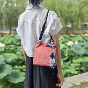 May include: A pink canvas tote bag with a black strap and a floral patterned charm. The bag has a single pocket on the front.