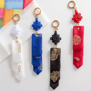 Op de afbeelding: Vier sleutelhangers met verschillende gekleurde stoffen kwastjes en goudkleurige knopen. De kwastjes zijn versierd met goudkleurige ronde patronen. De sleutelhangers zijn wit, blauw, zwart en rood.