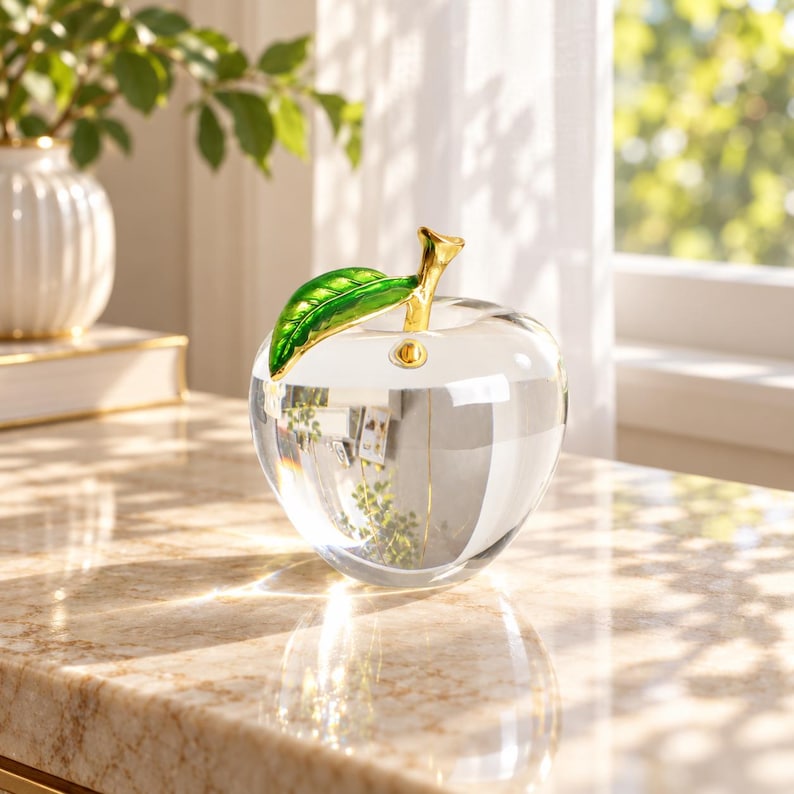 May include: A clear glass apple figurine featuring a polished gold-tone stem and a vibrant green enameled leaf. The apple has a smooth, rounded shape and sits as a decorative object, reflecting light through its transparent body.