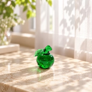 May include: A small, translucent emerald green glass apple figurine featuring a smooth, rounded body and a single detailed leaf attached to the stem. The glass material catches the light, creating subtle reflections and internal highlights.