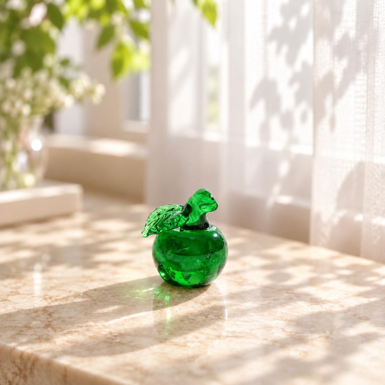 May include: A small, translucent emerald green glass apple figurine featuring a smooth, rounded body and a single detailed leaf attached to the stem. The glass material catches the light, creating subtle reflections and internal highlights.