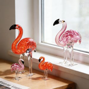 May include: Four glass flamingo ornaments of different sizes. The flamingos have clear glass legs and black-tipped beaks. The bodies are shades of pink and orange with speckled patterns. They are displayed on a wooden surface near a window.