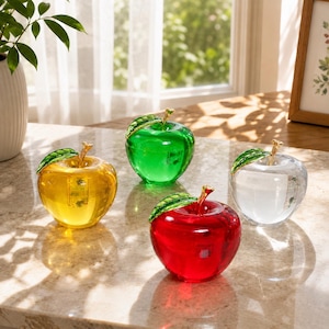 May include: Four decorative glass apples in yellow, green, red, and clear colors. Each apple features a gold-toned stem and a single green leaf attached to the top. The smooth, translucent surfaces reflect light, creating a glossy appearance.