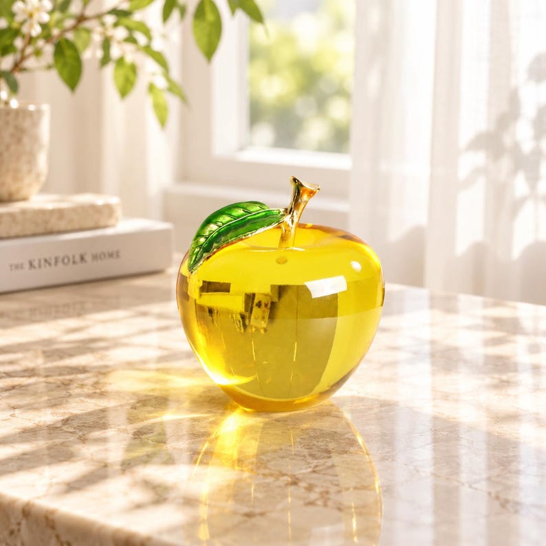 May include: A translucent yellow glass apple figurine featuring a smooth, rounded body and a metallic gold stem attached to a single, vibrant green glass leaf. The object reflects light, creating bright highlights and a clear, polished appearance.