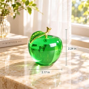 May include: A translucent green glass apple figurine featuring a polished gold-tone metal stem and leaf. The apple measures 2.17 inches in width and 2.24 inches in height, showcasing a smooth, reflective surface that catches the light.