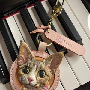 Peut inclure: Un porte-clés en forme de chat avec un dos en cuir rose et un fermoir doré. Le chat a de grands yeux expressifs et un collier de perles colorées qui épelle "PEARL". Une étiquette rose avec le mot "Pearl" est attachée.