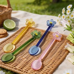 Könnte beinhalten: Vier farbige Glaslöffel mit blütenförmigen Verzierungen. Die Löffel sind gelb, grün, blau und rosa und liegen auf einem geflochtenen Tablett. Geeignet zum Servieren oder Rühren.