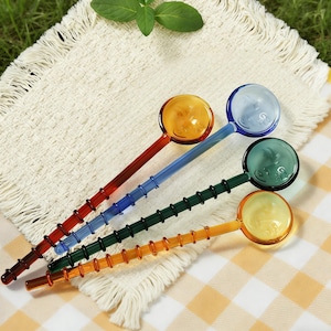 May include: Four colorful glass spoons with round bowls and long, textured handles. The spoons are in shades of red, blue, green, and orange. The spoons are arranged on a white fringed cloth and a yellow and white checkered cloth.