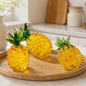 May include: Three pineapple-shaped glass ornaments featuring translucent yellow bodies with a textured diamond pattern and vibrant green glass leaves at the top. These decorative items are arranged on a round, woven rattan tray.