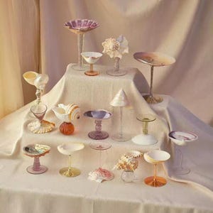 Puede incluir: Una colección de objetos decorativos de vidrio y conchas marinas, que incluyen copas con pie y esculturas de conchas marinas. Los artículos están dispuestos sobre un mantel blanco, con un fondo de tonos suaves y neutros. Los tallos de vidrio tienen varios colores y las conchas marinas son de tonos naturales.