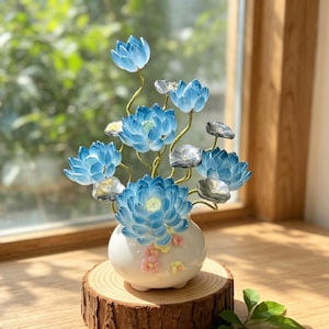 Op de afbeelding: Een decoratief bloemstuk met blauwe lotusbloemen in een witte keramische vaas. De vaas staat op een houten voet. De bloemen hebben een doorschijnende kwaliteit en het arrangement bevat grijze en gele accenten.