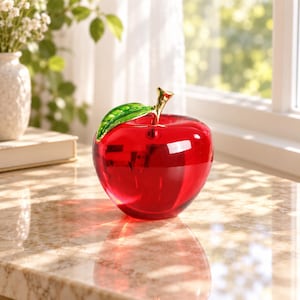 May include: A bright red glass apple figurine featuring a smooth, translucent surface. The apple is topped with a single, vibrant green glass leaf and a polished gold-tone metal stem, creating a reflective and polished decorative object.