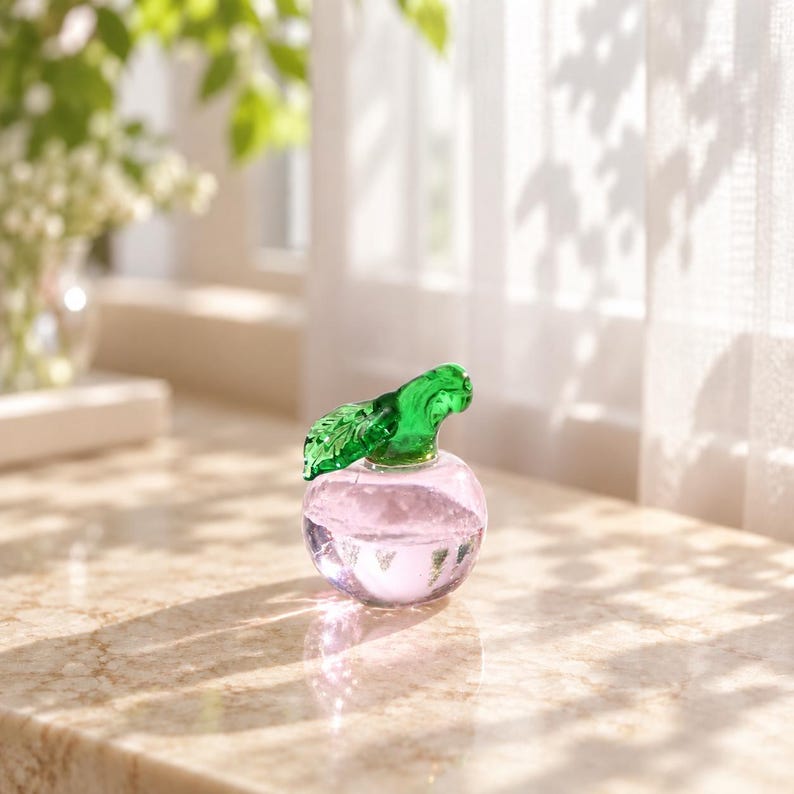 May include: A small, round, clear glass figurine shaped like an apple with a light pink tint in the base and a vibrant green glass leaf attached to the top stem. The object is smooth, translucent, and reflects light on its curved surface.