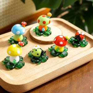May include: A collection of colorful glass figurines, including mushrooms and a ladybug, displayed on a wooden tray. The mushrooms feature red, yellow, blue, and green caps with white polka dots. Each figurine rests on a green, four-leaf clover base.