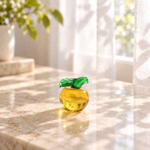 May include: A small, round, yellow glass apple figurine featuring a smooth, translucent body and a vibrant green glass leaf attached to the top stem. The object is crafted from clear yellow glass with subtle internal bubbles and a polished finish.