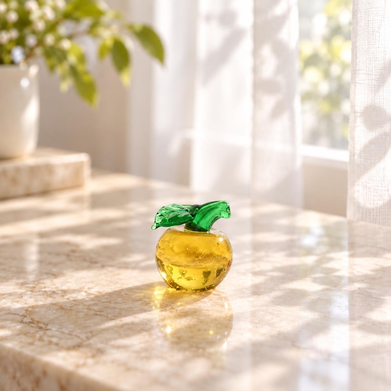 May include: A small, round, yellow glass apple figurine featuring a smooth, translucent body and a vibrant green glass leaf attached to the top stem. The object is crafted from clear yellow glass with subtle internal bubbles and a polished finish.