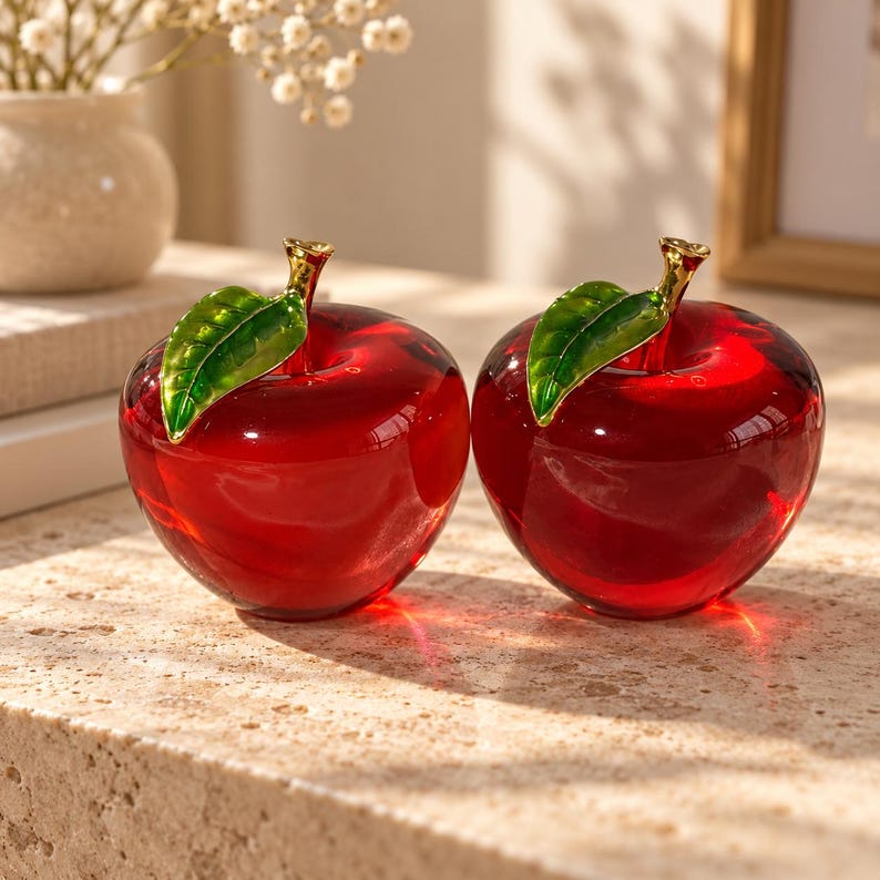 May include: Two translucent red glass apple figurines, each featuring a polished gold-tone stem and a single vibrant green enameled leaf attached to the top. The apples have a smooth, rounded shape and a glossy finish that reflects light.