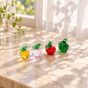 May include: Four small glass apple figurines arranged in a row. The first is yellow, the second is light pink, the third is bright red, and the fourth is green. Each apple features a small, detailed green glass leaf attached to its stem.