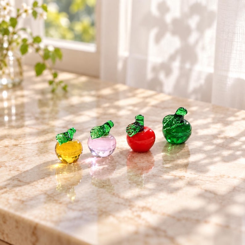 May include: Four small glass apple figurines arranged in a row. The first is yellow, the second is light pink, the third is bright red, and the fourth is green. Each apple features a small, detailed green glass leaf attached to its stem.