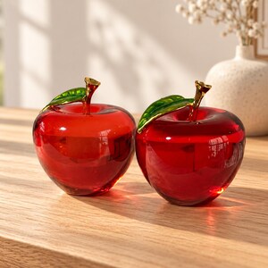 May include: Two red glass apple figurines, each featuring a smooth, translucent red body and a metallic gold stem topped with a single, glossy green leaf. The apples are crafted with a polished finish that reflects light, creating a clear, vibrant appearance.