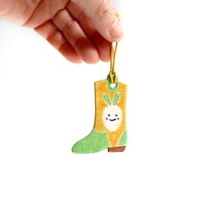 May include: A green and yellow ceramic boot ornament with a white smiling carrot design. The boot has a yellow top and a green bottom with a brown heel. The ornament is hanging from a yellow string.