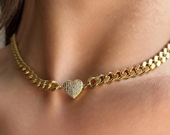 Może przedstawiać: Złoty naszyjnik typu choker z zawieszką w kształcie serca. Zawieszka jest inkrustowana małymi, błyszczącymi kamieniami. Naszyjnik ma wzór łańcucha typu curb, z zapięciem. Biżuteria jest noszona wokół szyi.