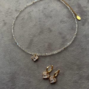 May include: A delicate gold necklace with a clear crystal bead chain and a small heart pendant. Matching heart-shaped earrings with clear crystals dangle from gold hoops.