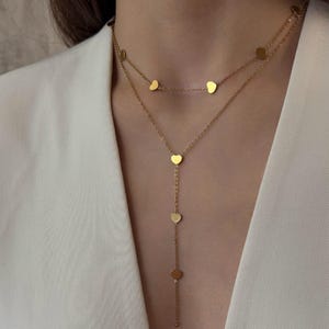 May include: A gold-colored necklace featuring a layered design. The top layer has small heart-shaped pendants, while the lower layer extends into a Y-shape with additional heart pendants. The necklace is worn against a white blazer.