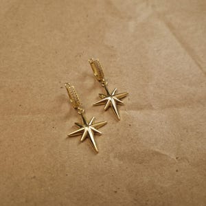 May include: A pair of gold-colored starburst earrings. Each earring features a star-shaped pendant suspended from a hoop. The hoops and star pendants have a shiny, metallic finish. The earrings are displayed on a brown surface.