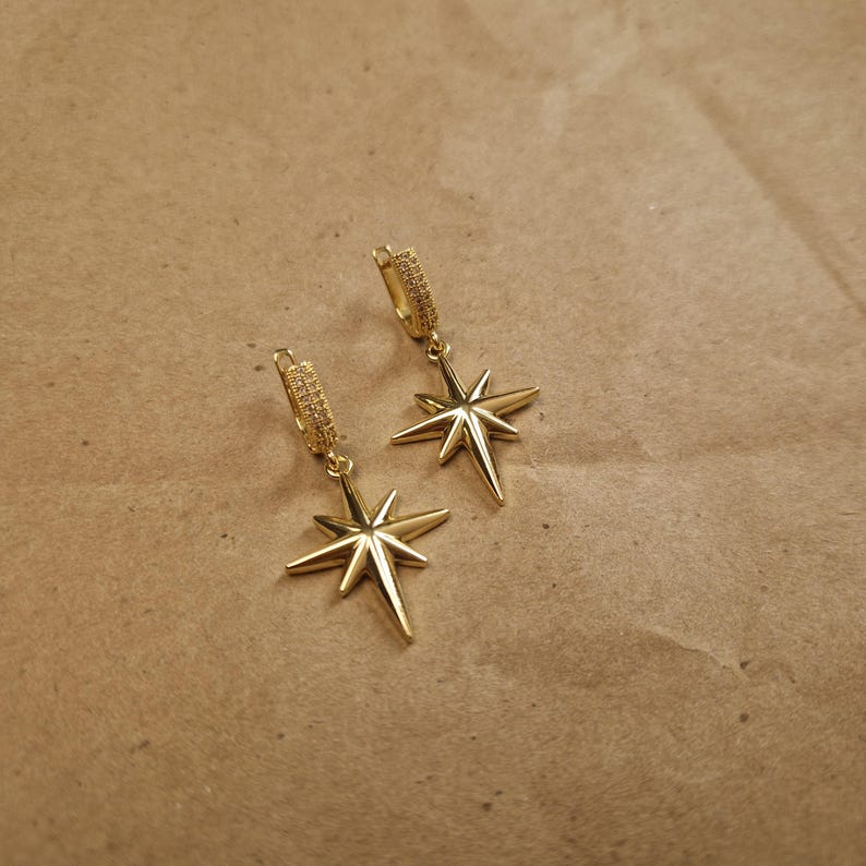 May include: A pair of gold-colored starburst earrings. Each earring features a star-shaped pendant suspended from a hoop. The hoops and star pendants have a shiny, metallic finish. The earrings are displayed on a brown surface.