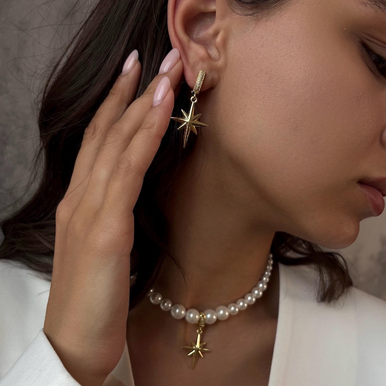May include: Gold-toned starburst earrings and a matching pendant necklace with a pearl strand. The earrings feature a small hoop with a starburst charm. The necklace has a similar starburst pendant. The jewelry is worn with a white blazer.