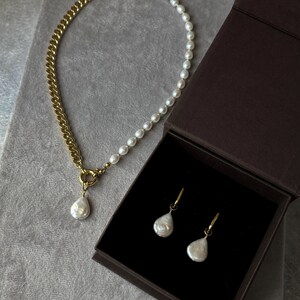 Puede incluir: Un collar de cadena dorada y perlas con un colgante de perla en forma de lágrima. Pendientes de perlas a juego con aros dorados se exhiben en una caja de joyería marrón. La joyería se encuentra sobre un fondo gris.