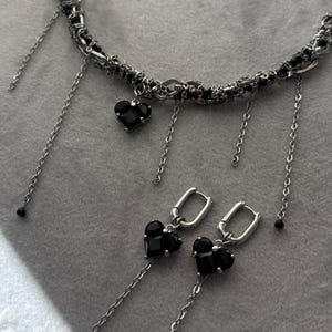K&ouml;nnte beinhalten: Eine silberne Kettenkette mit schwarzen herzf&ouml;rmigen Anh&auml;ngern und passenden Ohrringen. Die Kette hat mehrere Kettenstr&auml;nge und die Ohrringe haben ein Ringdesign.