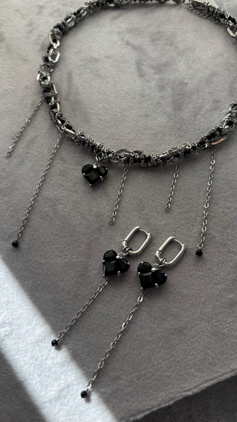 K&ouml;nnte beinhalten: Eine silberne Kettenkette mit schwarzen herzf&ouml;rmigen Anh&auml;ngern und passenden Ohrringen. Die Kette hat mehrere Kettenstr&auml;nge und die Ohrringe haben ein Ringdesign.