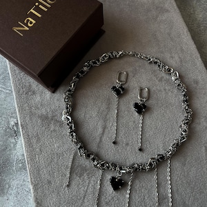 K&ouml;nnte beinhalten: Eine silberne Kettenkette mit schwarzen Herzanh&auml;ngern und passenden Ohrringen. Die Kette hat mehrere baumelnde Ketten mit schwarzen Perlen an den Enden. Der Schmuck ist in einer braunen Schachtel mit dem Text "NaTilo" auf der Oberseite verpackt.