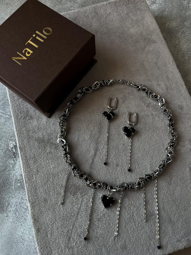 K&ouml;nnte beinhalten: Eine silberne Kettenkette mit schwarzen Herzanh&auml;ngern und passenden Ohrringen. Die Kette hat mehrere baumelnde Ketten mit schwarzen Perlen an den Enden. Der Schmuck ist in einer braunen Schachtel mit dem Text "NaTilo" auf der Oberseite verpackt.
