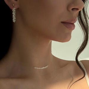 May include: A delicate jewelry set featuring a pair of dangling earrings and a necklace. The earrings and necklace are adorned with small, clear beads, creating a sparkling effect. The earrings hang from gold-colored posts, and the necklace appears to float on the skin.