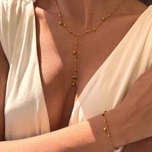 May include: A gold-colored necklace and bracelet set. The necklace features a delicate chain with small, evenly spaced beads and a longer, beaded drop. The bracelet mirrors the necklace design. The jewelry is displayed against a white background.