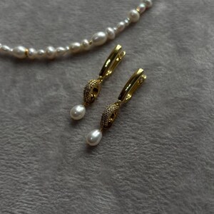 May include: A pair of gold hoop earrings with a pearl drop and a small crystal design. The earrings are on a white fuzzy surface.