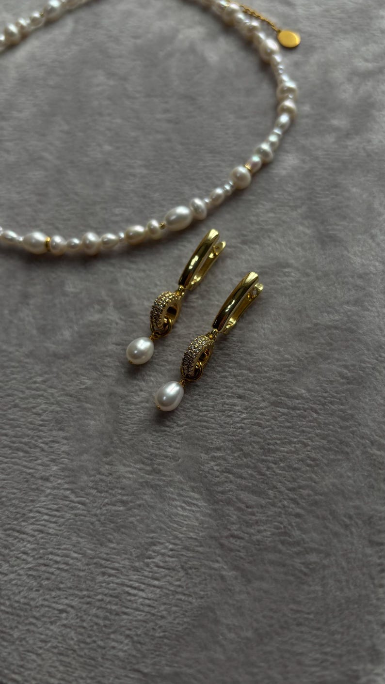 May include: A pair of gold hoop earrings with a pearl drop and a small crystal design. The earrings are on a white fuzzy surface.