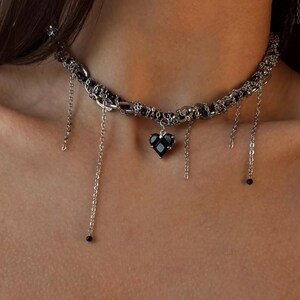 K&ouml;nnte beinhalten: Eine silberne Choker-Kette mit mehreren Ketten und einem schwarzen Herz-Anh&auml;nger. Die Ketten haben kleine schwarze Perlen an den Enden.
