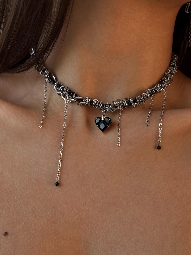 K&ouml;nnte beinhalten: Eine silberne Choker-Kette mit mehreren Ketten und einem schwarzen Herz-Anh&auml;nger. Die Ketten haben kleine schwarze Perlen an den Enden.