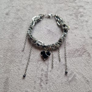 May include: Silver bracelet featuring a woven design and dangling chains. A black heart-shaped charm is suspended from the centre. The bracelet is on a grey surface.