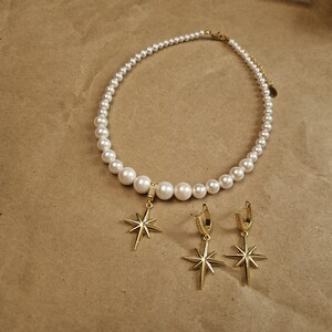 May include: A pearl necklace and matching earrings set. The necklace features white pearls of varying sizes, with a gold-colored clasp. The earrings and pendant are gold-colored starburst designs, with small clear stones.