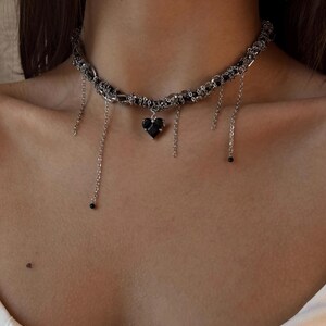 K&ouml;nnte beinhalten: Eine silberne Choker-Kette mit einem schwarzen Herz-Anh&auml;nger und herabh&auml;ngenden Ketten. Die Kette besteht aus mehreren miteinander verflochtenen Ketten.