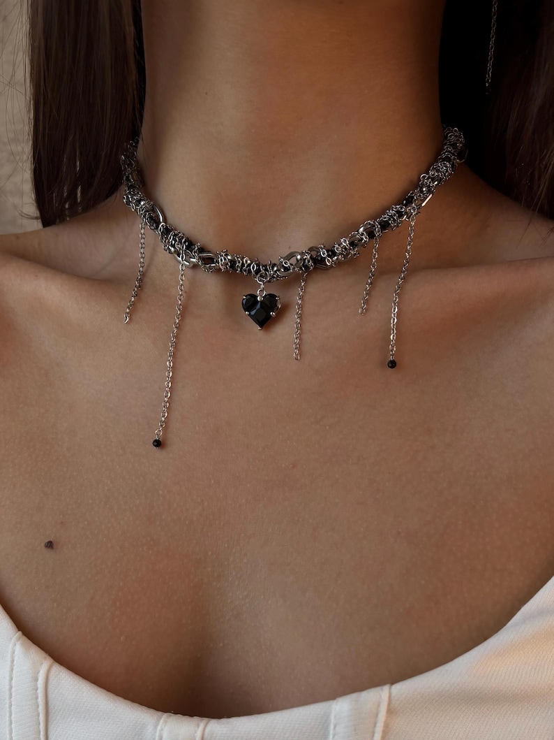 K&ouml;nnte beinhalten: Eine silberne Choker-Kette mit einem schwarzen Herz-Anh&auml;nger und herabh&auml;ngenden Ketten. Die Kette besteht aus mehreren miteinander verflochtenen Ketten.