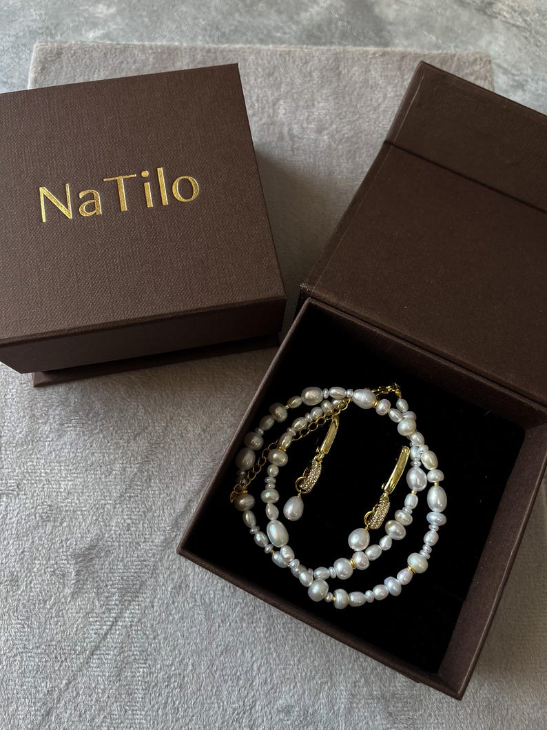 May include: A set of pearl earrings and a pearl necklace in a brown box with the text "Na Tilo" on the lid.